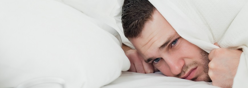 En ung man ligger i sin säng och håller sitt täcke över sitt huvud och tittar missnöjt ned mot klockan som om han inte kan sova på grund av störande ljud.