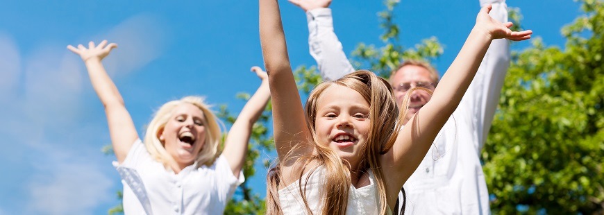 En glad tjej hoppar upp och sträcker sina händer i luften en varm sommardag samtidigt som hennes mamma och pappa som står bakom henne gör detsamma. I bakgrunden syns en blå sommarhimmel och siluetten av ett frisktgrönt träd. 