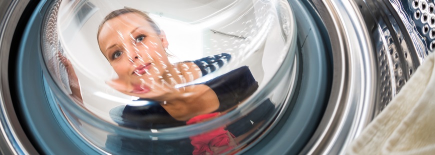 Från insidan av en tvättmaskin ser man att en medelålders kvinna sitter på knä utanför maskinen och tittar in genom den välvda glasrutan där tvätten ligger.
