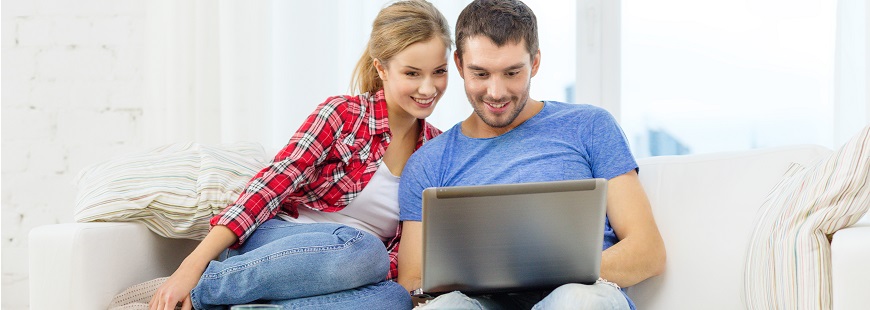 Ett leende ungt par sitter i en soffa och kollar på skärmen till en laptop.
