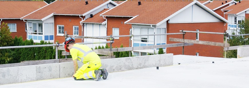 En byggnadsarbetare ligger på knä och mäter med tumstock på ett nygjutet våningsplan. Bakom en byggnadsställning ses huskropparna från ett närbeläget kvarter av hyreshus.