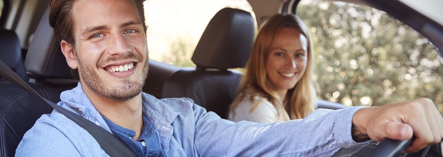 I en bil med nerrullad sidoruta sitter en medelålders man och en medelålders kvinna och ler mot kameran, mannen håller sin högra hand över ratten. 