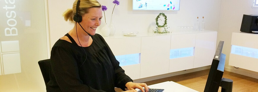 En kvinnlig kundservicemedarbetare sitter vid en dator i en ljus och öppen reception. Kvinnan har ett headset på sig när hon leende skriver på datorns tangentbord.