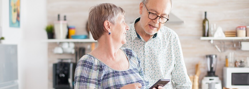 Ett pensionärspar står i sitt kök och kvinnan låter maken titta på skärmen till en smartphone som hon håller i sin hand.