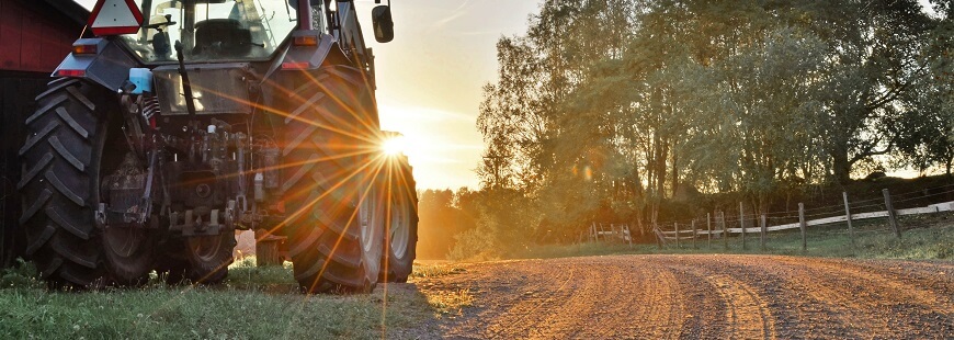 Invid en grusväg ute på landet står en traktor. Solen kastar sina strålar över traktorns hjul och bländar kameran.