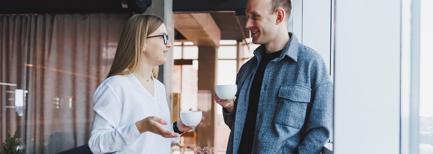 En man och en kvinna står i ett kontorslandskap och samtalar över en kopp kaffe. 