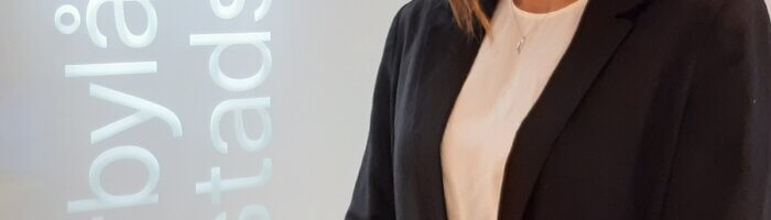 En kvinna i mörk kostymjacka står framför en glasskärm med MBABs logotyp i vitt.